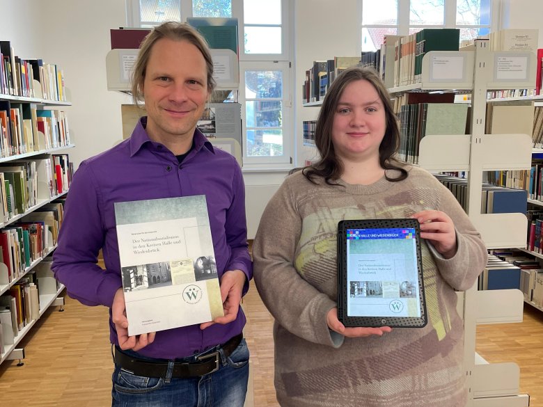 EIn Mann hält ein Buch in der Hand. Eine Frau ein Tablet, auf dem eine App geöffnet ist. Im Hintergrund sind Bücherregale im Archiv zu sehen. 