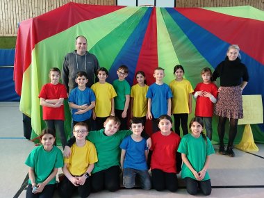 Kinder tragen bunte T-Shirts, stehen nebeneinander. Zwei erwachsene Personen stehen link und rechts von ihnen. 