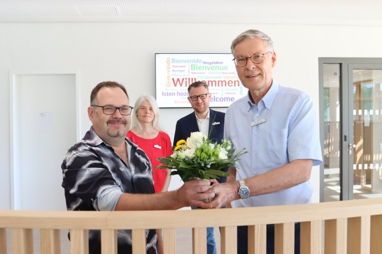 2 Personen im Vordergrund, wobei die eine der anderen einen Blumenstrauß überreicht. Im Hintergrund stehen zwei weitere Personen,