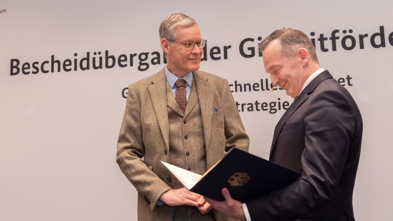 Zwei Männer stehen nebeneinander. Eine Person hält den Bescheid in der Hand.