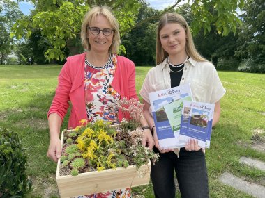 Zwei Frauen stehen im Grünen und halten Infomaterial sowie eine Box zur Darstellung einer Dachbegrünung in den Händen.