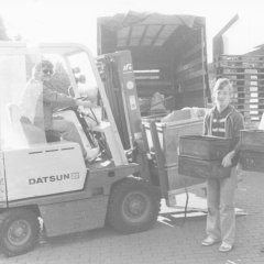 Schwarz-weiß-Foto: Ein Mensch im Gabelstapler und zwei mit Kisten in der Hand beladen einen Transporter.