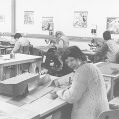 Schwarz-Weiß-Foto: Eine Frau mit einem Stapel Stoffe auf dem Tisch lächelt in die Kamera. Im Hintergrund sind weitere Menschen an Nähmaschinen zu sehen.