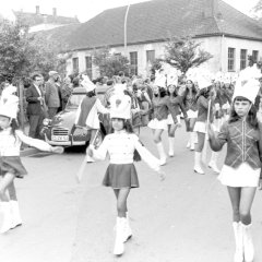 Schwarz-Weiß-Foto: Eine Gruppe Frauen und Kinder mit Uniformen ziehen an einer Gruppe zuschauender Menschen vorbei. 